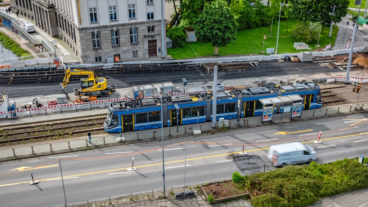 Das Bild zeigt eine Skoda-Straßenbahn auf den Gleisen am Falkeplatz. Hinter der Bahn ist die Baustelle sowie ein gelber Bagger erkennbar.